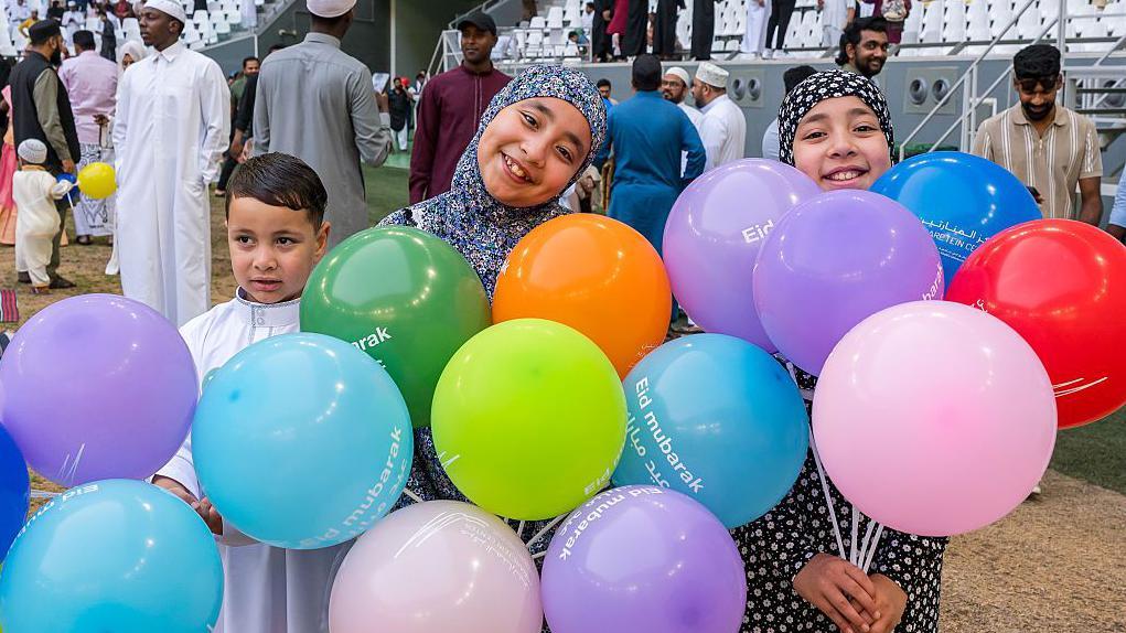 ثلاثة أطفال يحملون بالونات بعدة ألوان بعد أداء صلاة عيد الأضحى المبارك في استاد المدينة التعليمية بالدوحة، قطر، في أول أيام العيد، في السادس من يونيو/حزيران 2025.