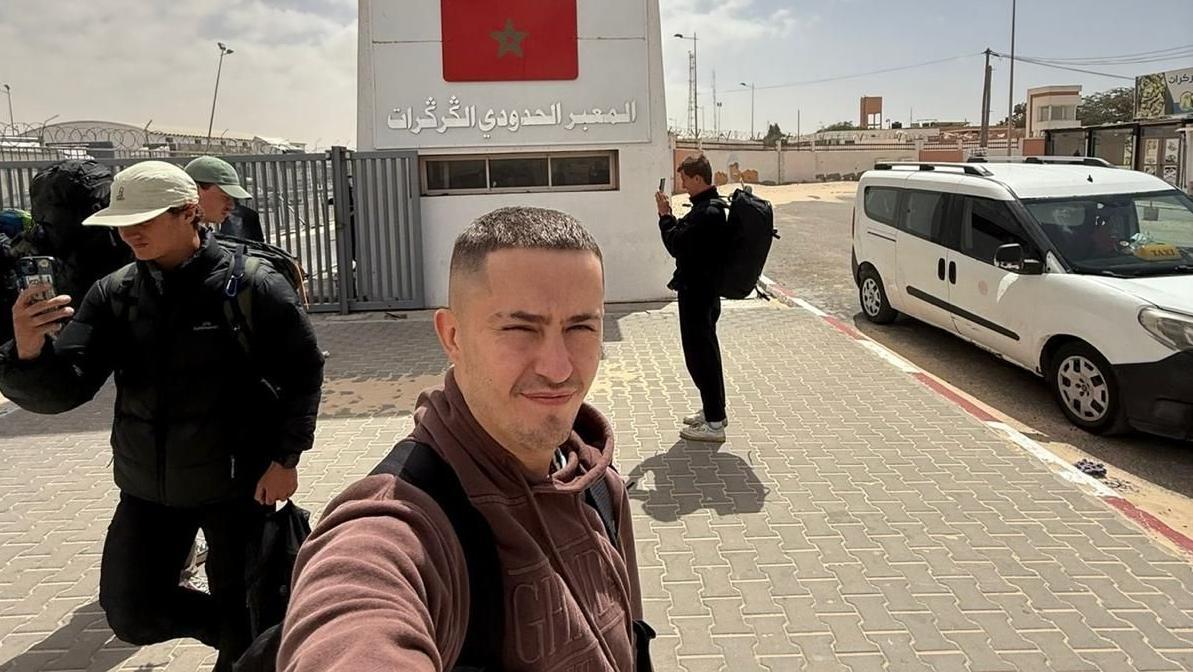 توم روك يلتقط صورة سيلفي أمام معبر الكركرات الحدودي بين الصحراء الغربية وموريتانيا. وخلفه هيكل خرساني ضخم مرسوم عليه العلم المغربي.